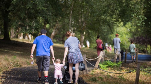 Mother and father holding their child's hands walking along a tarmacked pathway at Winkworth Arboretum. Mum is also pushing a pram. There are other people walking in the background.